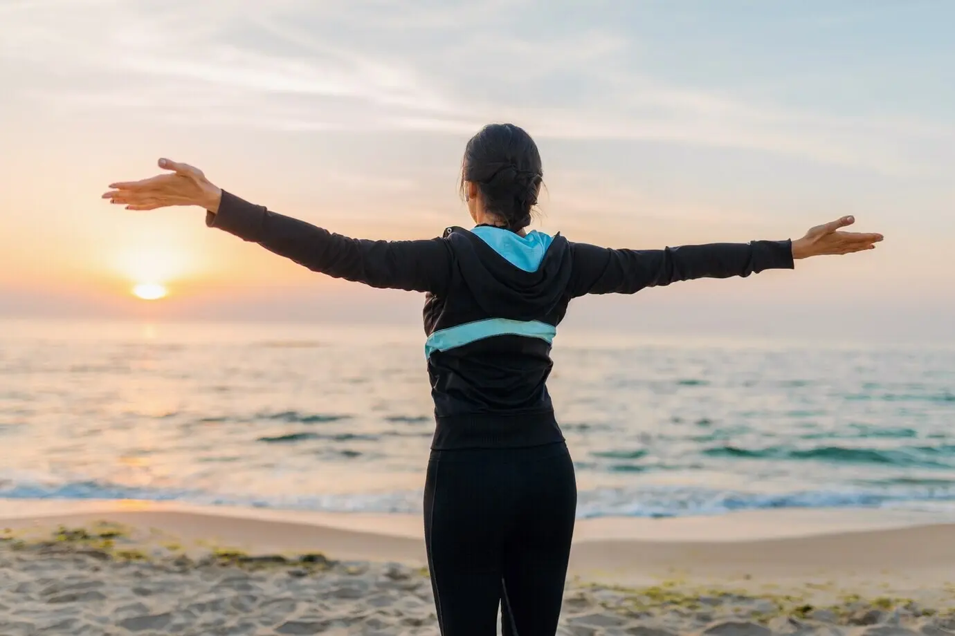 A young, attractive, slim woman, viewed from behind, greeting the sun, holding hands, doing sport exercises on a beach at morning sunrise, in sportswear, healthy lifestyle.